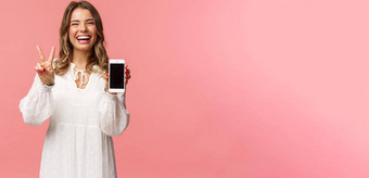 肖像卡哇伊乐观快乐年轻的女孩白色衣服显示移动电话显示和平标志笑感觉快乐的很高兴分享太棒了应用程序链接粉红色的背景