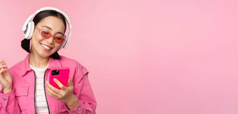 时尚的亚洲女孩跳舞智能<strong>手机</strong>听音乐耳机移动电话应用程序微笑笑摆姿势粉红色的背景