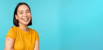 疫苗运动科维德快乐健康的亚洲女孩笑疫苗接种冠状病毒邦迪牌创可贴肩膀穿黄色的t恤蓝色的背景