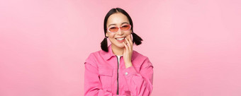 关闭肖像时尚的亚洲女人太阳镜微笑受宠若惊笑卖弄风情的站粉红色的背景