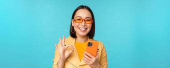 快乐朝鲜文女孩太阳镜显示标志持有移动电话智能<strong>手机</strong>应用程序推荐应用程序站蓝色的背景