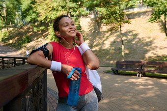 年轻的女运动员运动服微笑美丽的露出牙齿的微笑持有瓶水把耳机放松早....慢跑功能有氧运动锻炼城市公园