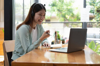 微笑女孩学生穿无线耳机研究在线老师快乐年轻的亚洲女人学习语言听讲座看网络研讨会写笔记移动PC坐咖啡馆遥远的教育