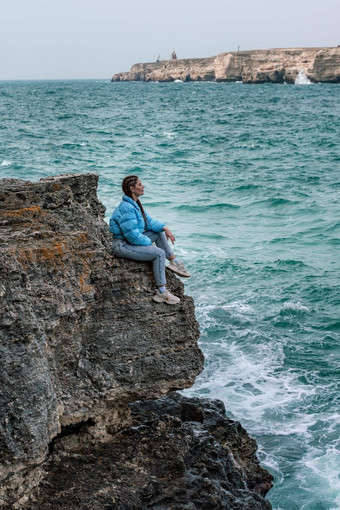 女人蓝色的夹克坐在岩石悬崖海狂风暴雨的海洋女孩旅行者休息认<strong>为梦想</strong>享受自然和平平静景观多风的天气