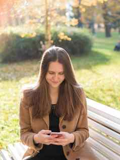 美丽的青少年女孩坐着公园板凳上持有智能手机聊天在线互联网女人长头发