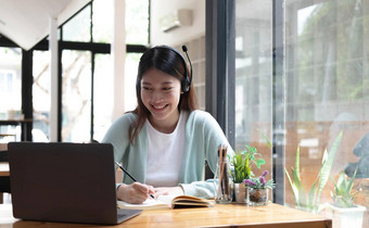 微笑女孩学生穿无线耳机研究在线Skype老师快乐年轻的女人学习语言听讲座看网络研讨会写笔记移动PC坐咖啡馆遥远的教育