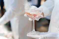 服务员白色手套持有酒玻璃闪闪发光的酒红色的樱桃白色烟干冰客户