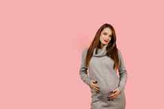 怀孕了女人期望孩子孕妇离开怀孕快乐美丽的女孩灰色的毛衣粉红色的背景