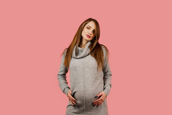怀孕快乐美丽的女孩灰色的毛衣粉红色的背景怀孕了女人期望孩子孕妇离开