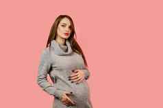 怀孕快乐美丽的女孩灰色的毛衣粉红色的背景怀孕了女人期望孩子孕妇离开
