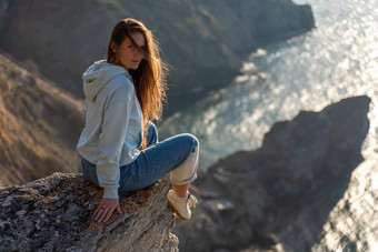 女人<strong>旅游</strong>享受日落海山景观坐在在<strong>户外</strong>岩石海穿牛仔裤蓝色的连帽衫健康的生活方式和谐冥想