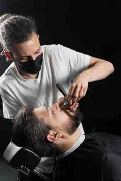 胡子剃须理发店理发师长发绺黑色的医疗面具修剪胡子英俊的男人。检疫冠状病毒科维德