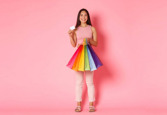 完整的长度肖像快乐时尚的亚洲女孩显示折扣信贷卡持有购物袋享受<strong>花钱</strong>衣服站粉红色的背景