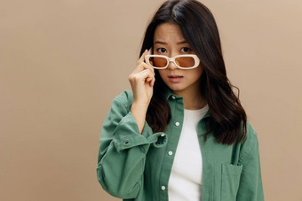 特写镜头肖像可爱的朝鲜文年轻的女人卡其色绿色衬衫触摸时尚的眼镜相机摆姿势孤立的米色柔和的工作室背景很酷的时尚提供太阳镜概念