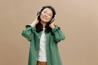放松享受最喜欢首歌可爱的亚洲学生年轻的女人卡其色绿色衬衫触摸耳机摆姿势孤立的米色<strong>柔和</strong>的工作室背景很酷的时尚提供<strong>音乐</strong>应用程序平台概念