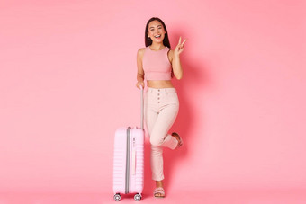 旅行假期假期概念快乐的亚洲女孩夏天衣服包装袋旅行在国外站手提箱微笑显示卡哇伊和平手势粉红色的背景