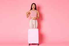 旅行假期假期概念全身的好奇的感兴趣亚洲旅游女孩夏天衣服持有手提箱指出手指想知道粉红色的背景