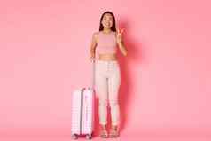 旅行假期假期概念可爱的亚洲女孩准备好了探索国家显示和平卡哇伊标志微笑旅行在国外旅游站手提箱粉红色的背景