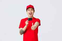兴奋微笑亚洲交付的家伙红色的帽t恤统一的显示翘拇指信贷卡推荐客户端伟大的提供促销快递服务在线购物互联网商店概念