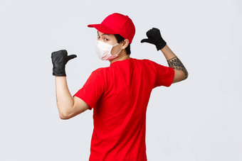 回来视图亚洲交付男人。医疗面具保护手套穿红色的帽t恤转相机指出回来显示公司标志统一的物流快递购物概念