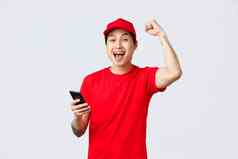 快乐快乐的亚洲交付的家伙红色的统一的帽t恤航空公司服务持有智能手机阅读好新闻高喊拳头泵大喊庆祝胜利achivement
