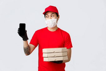 有趣的交付的家伙红色的帽t恤持有披萨盒子显示<strong>智能</strong>手机屏幕亚洲快递确认订单信息客户端带食物人首页检疫穿<strong>医疗</strong>面具