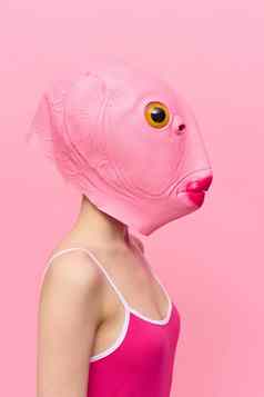 女人粉红色的鱼头面具站配置文件粉红色的背景相机黄色的眼睛疯狂的概念上的万圣节服装