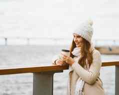 美丽的年轻的女孩喝咖啡茶塑料杯子秋天冬天女人长头发站海滨波罗的海海港口等待渡船加热热喝复制空间