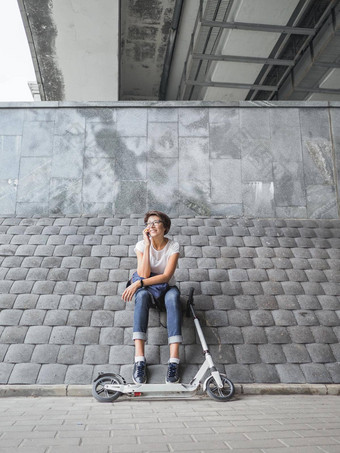 微笑女人会谈智能手机骑踢踏板车女牛仔裤运动鞋灰色平铺的墙背景健康的生活方式环保城市运输
