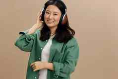 享受声音快乐可爱的亚洲学生年轻的女人卡其色绿色衬衫触摸耳机摆姿势孤立的米色柔和的工作室背景很酷的时尚提供音乐应用程序平台概念