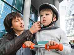 父亲紧固带儿子的头盔孩子培训骑踢踏板车道德支持城市生活方式