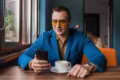 时尚的的家伙商人欧洲外观太阳镜夹克衬衫坐在表格咖啡馆咖啡打破持有智能手机移动电话手