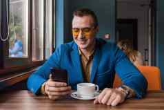 时尚的的家伙商人欧洲外观太阳镜夹克衬衫坐在表格咖啡馆咖啡打破持有智能手机移动电话手