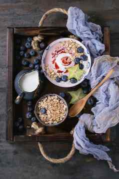 奶昔碗健康的早餐草莓酸奶蓝莓格兰诺拉麦片薄荷壶牛奶杨桃木托盘纺织纱布黑暗金属纹理背景前视图