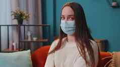 肖像年轻的生病的女孩穿医疗保护面具坐着生活房间相机