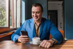 时尚的快乐的家伙商人欧洲外观夹克衬衫坐在表格咖啡馆咖啡打破持有智能手机移动电话手