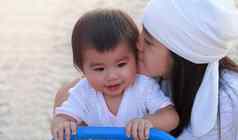 关闭肖像美丽的年轻的亚洲妈妈。接吻婴儿公园快乐可爱的妈妈。吻女儿微笑女孩玩操场上妈妈。
