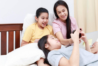 快乐亚洲家庭享受放松床上卧室妈妈。女儿享受平板电脑床上家庭概念