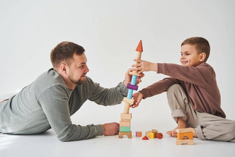 有爱心的爸爸帮助儿子玩地板上白色背景父亲孩子构建塔色彩斑斓的木砖有趣的