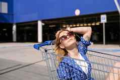 快乐的时尚的年轻的女人蓝色的衣服阳光明媚的一天坐在购物电车前面超市时尚很酷的微笑女孩