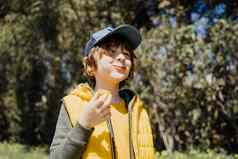 微笑快乐的孩子孩子黄色的背心绿色连帽衫吃脆零食在户外公共公园小学生男孩享受消耗咀嚼垃圾食物树植被背景