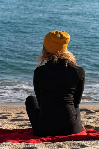 女孩金发碧眼的卷曲的头发黑色的体育<strong>运动夹克</strong>黄色的帽坐着海滩蓝色的海