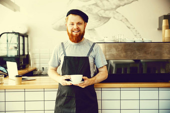 咖啡业务老板概念肖像快乐年轻的有胡子的高加索人咖啡师围裙自信微笑相机咖啡商店计数器