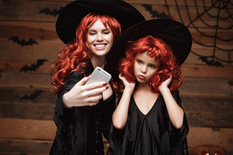 万圣节概念美丽的高加索人妈妈。女儿长红色的头发女巫服装采取自拍智能手机准备好了庆祝万圣节