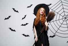 万圣节女巫概念肖像美丽的年轻的女巫飞天扫帚灰色墙蝙蝠蜘蛛网络背景
