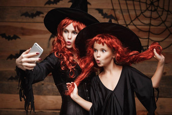 万圣节概念美丽的高加索人妈妈。女儿长红色的头发女巫服装采取自拍智能手机准备好了庆祝万圣节