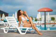 美丽的年轻的女人假期吃西瓜在游泳池边热一天