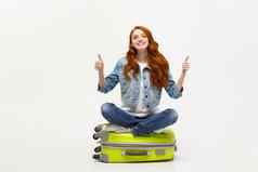 旅行生活方式概念年轻的兴奋高加索人女人坐着行李手提箱显示拇指孤立的白色准备好了假期
