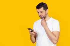 年轻的男人。胡子白色t恤智能手机自信表达式聪明的脸思考站孤立的黄色的背景