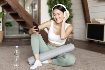 快乐运动女孩坐着地板上首页采取自拍锻炼穿耳机持有智能手机
